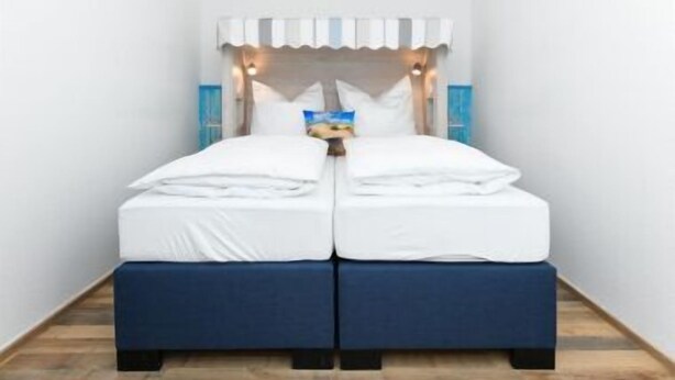 A double bed with white bedding and a blue headboard, set against a white wall with a window. - House Strandkorb apartment Strandkob 6 (Bensersiel)