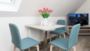 Una camera moderna con soffitto spiovente, un tavolino con un vaso di fiori, due sedie color petrolio e un televisore su un mobile.