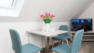 Una habitación moderna con techo inclinado, una pequeña mesa con un jarrón de flores, dos sillas color turquesa y un televisor sobre un mueble.