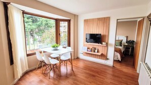 Ein modernes Zimmer mit Holzboden, einem weißen Tisch mit Stühlen, einem an einer Holzvertäfelung montierten Fernseher und Blick ins Grüne.