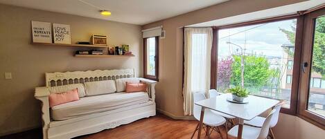 A cozy room with a white sofa, a dining table with chairs, and a window with a view of greenery.