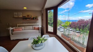 Ein gemütliches Zimmer mit einem weißen Sofa, einem Esstisch mit Topfpflanze und einem großen Fenster mit Blick auf eine Wohnstraße und Grünflächen.
