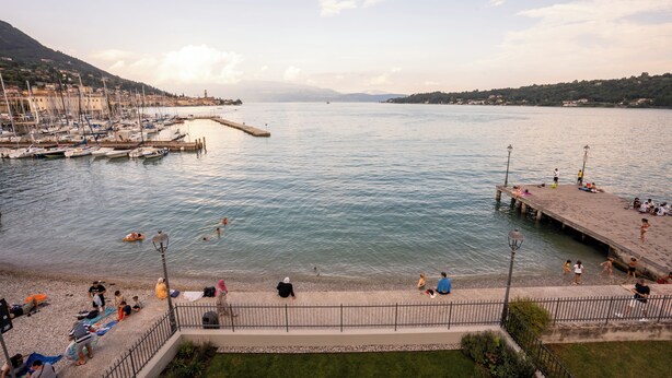 A marina with boats docked, a pier with people, and a view of the water and distant hills. - Apartment with a balcony overlooking the harbor (Salò)