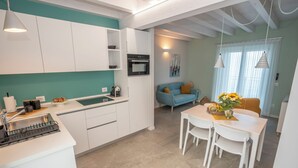 A modern kitchen with white cabinets, a built-in oven, and a sink. There is a dining area with a white table and chairs, and a blue sofa in the background. - Apartment with a balcony overlooking the harbor (Salò)