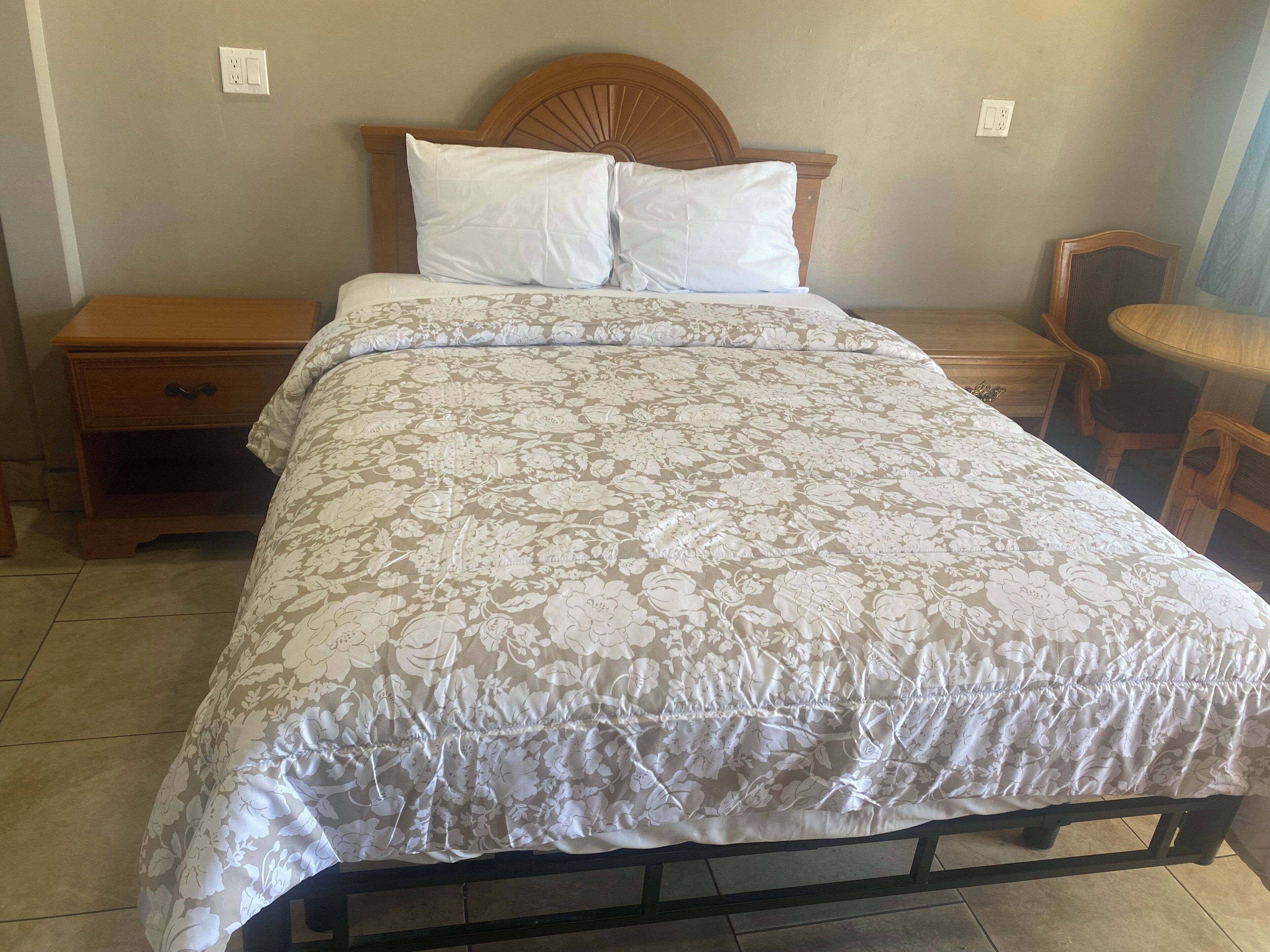A neatly made bed with a floral-patterned bedspread and two pillows, a wooden nightstand with a drawer, and a chair in the corner.