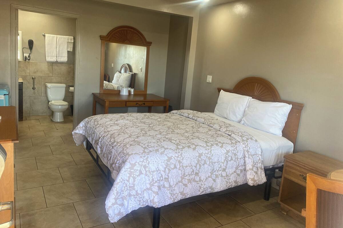 Un dormitorio con cama, una mesita de noche de madera, cómoda, espejo y baño con inodoro y toallero.