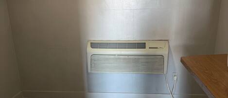 A room with a wooden floor, a white wall, and a white air conditioning unit mounted on the wall.