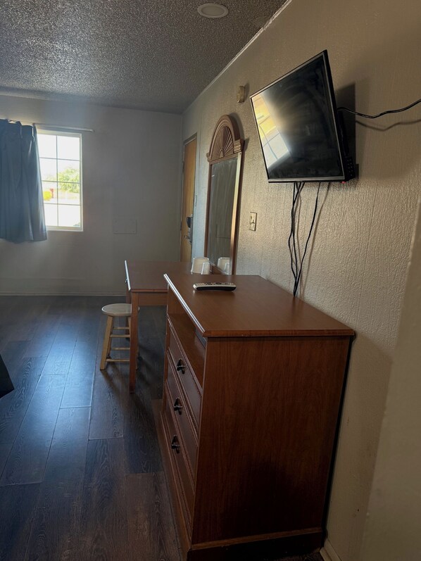 Una habitación con cómoda de madera, televisor montado en la pared, ventana con cortinas y suelo de madera.