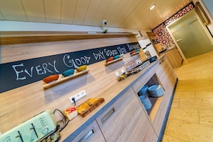 Uma área de recepção de hotel moderna com um balcão de madeira, uma lousa com o texto "EVERY GOOD DAY STARTS HERE" e diversos detalhes como uma balança, uma pequena planta e pratos empilhados de forma organizada.