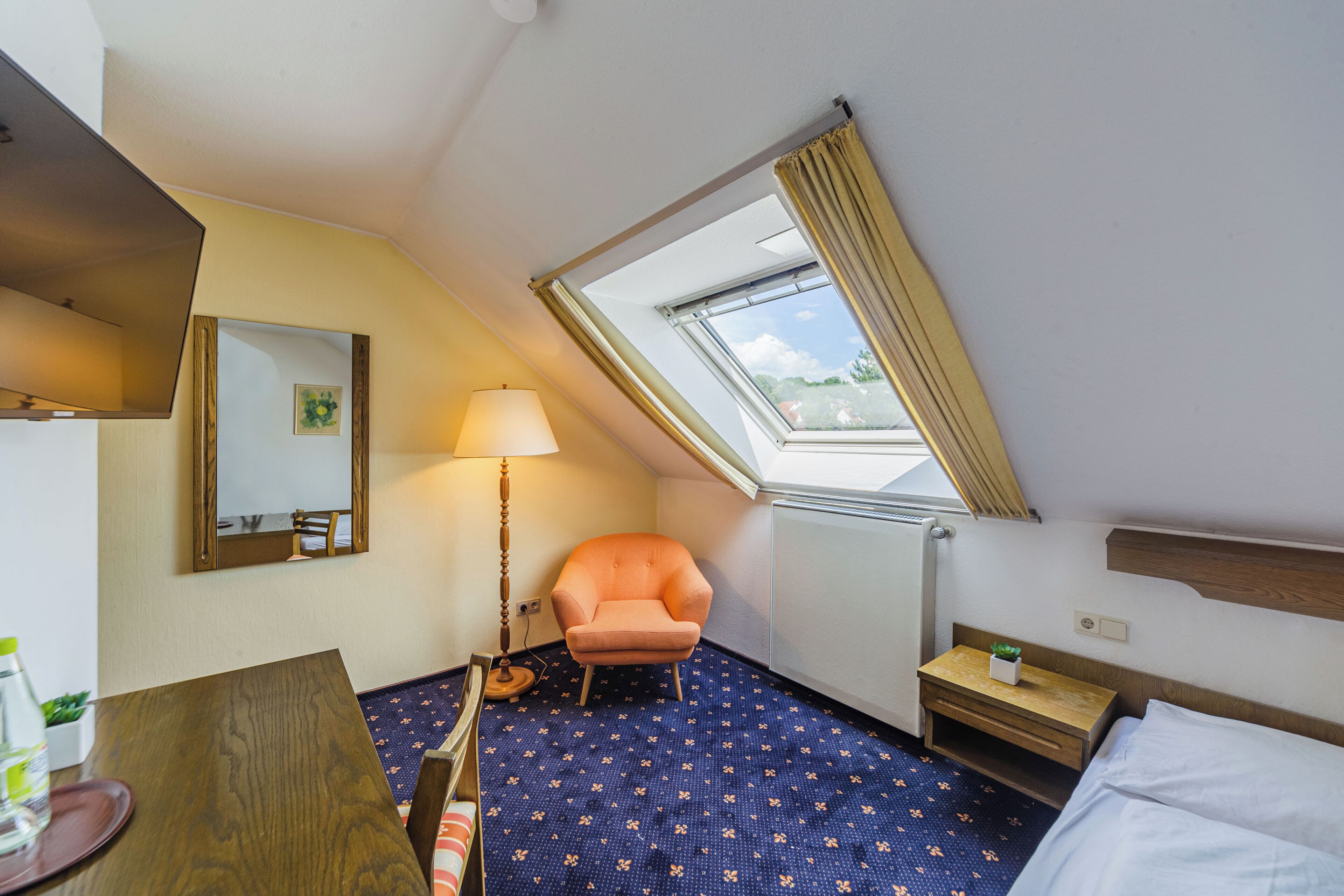 A hotel room with a bed, a chair, a table, a lamp, a small bedside table, and a window with curtains.