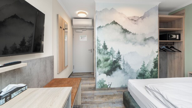 A hotel room with a bed, a wooden bench, a TV, a wardrobe, and a wall with a mountain and forest mural. - Hotel Franken (Forchheim)