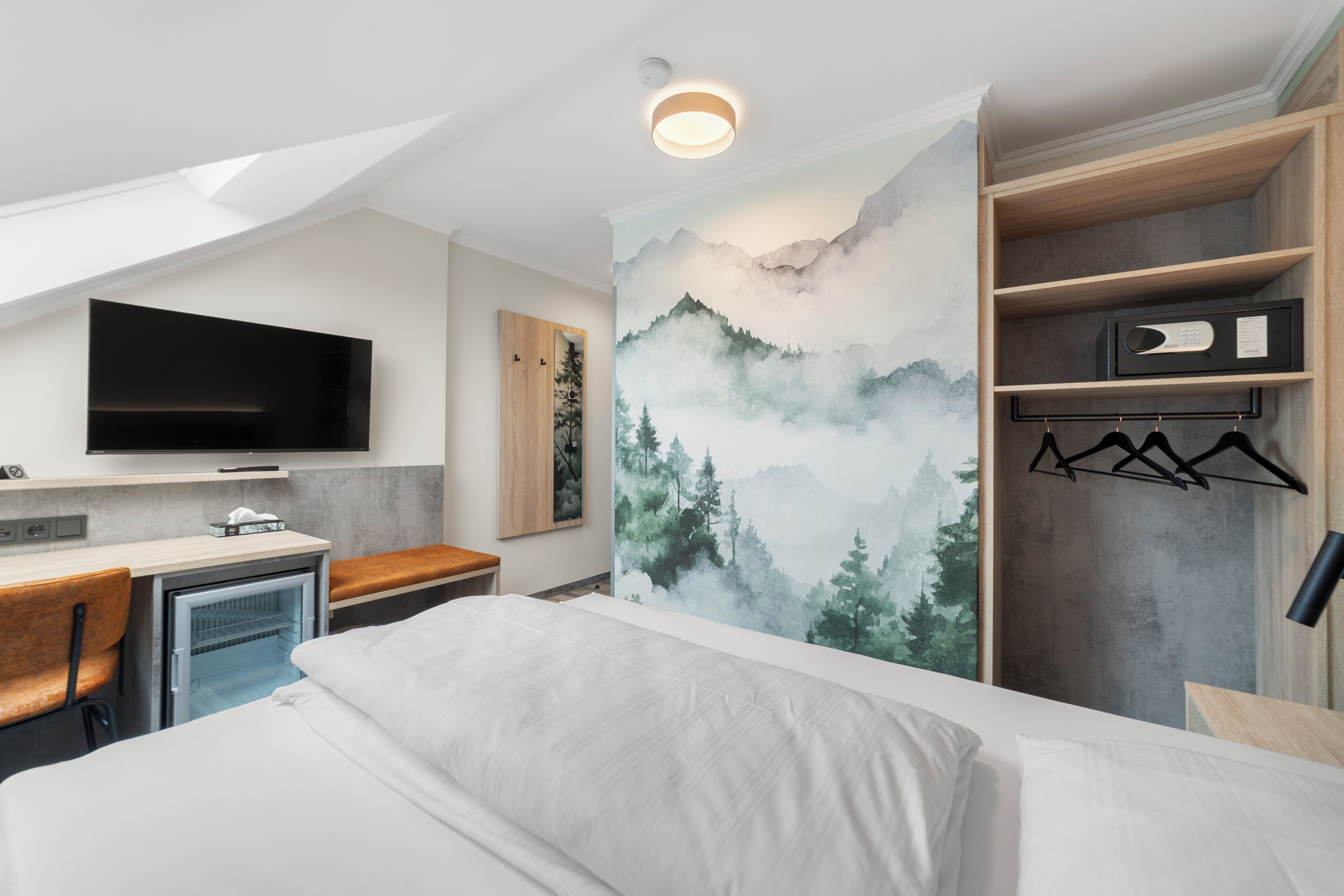 Ein modernes Hotelzimmer mit Bett, Schreibtisch mit Computer, Fernseher, Minikühlschrank und Kleiderschrank.