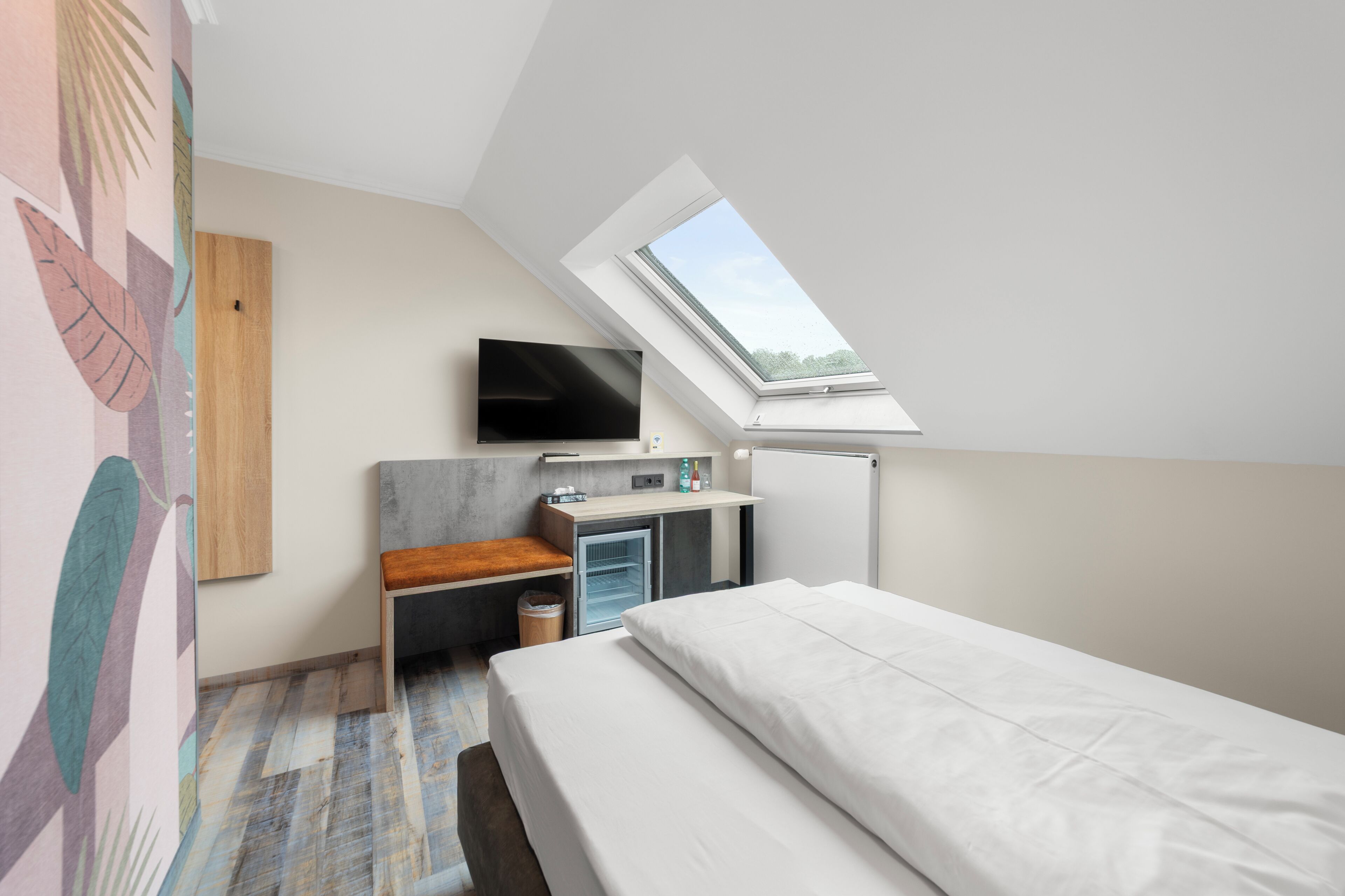 A bedroom with a skylight, a bed, a TV, a desk, and a small fridge.