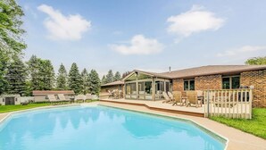 A swimming pool with a patio area, a covered seating area, and a brick building with large windows. - The Poolside Escape | Hot Tub | Arcade | Ping Pong (Bridgman)
