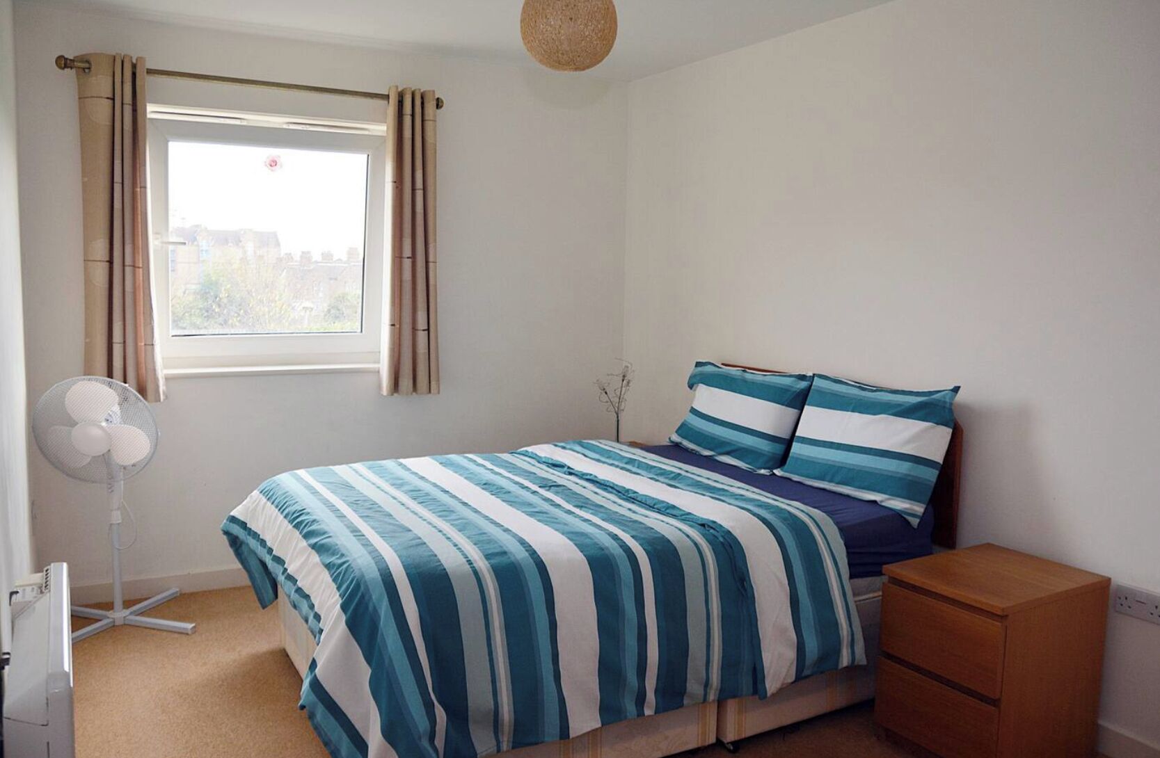 Une chambre à coucher comprenant un lit, une table de chevet, une commode, un ventilateur sur pied et une fenêtre avec des rideaux.
