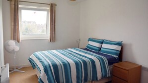 Ein Schlafzimmer mit einem Bett, einem Nachttisch, einem Ventilator und einem Fenster mit Vorhängen.