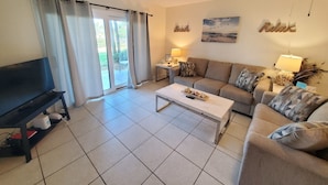 A living room with a sofa, coffee table, TV, and a view through a sliding door to the outdoors. - Ocean Village Club O15 (St. Augustine)