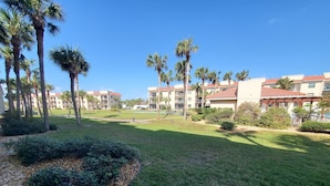 A well-maintained grassy area with palm trees and shrubs, surrounded by multi-story buildings with red-tiled roofs. - Ocean Village Club O15 (St. Augustine)
