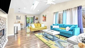 Ein geräumiges Wohnzimmer mit gelbem Sofa, blauem Sofa und weißem Couchtisch. Es gibt einen Deckenventilator, einen Kamin und einen Blick ins Freie durch große Fenster.