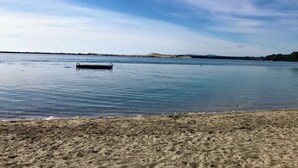 Ein Strand mit ruhiger See, einem kleinen Steg und sanften Hügeln im Hintergrund.