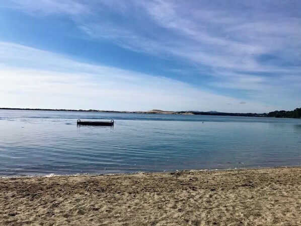 Ein Strand mit ruhiger See, einem kleinen Steg und sanften Hügeln im Hintergrund.