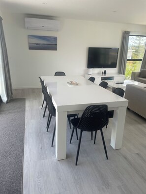 Ein moderner Essbereich mit einem weißen Tisch und schwarzen Stühlen, einem an der Wand montierten Flachbildfernseher und Blick auf Grünflächen außerhalb.