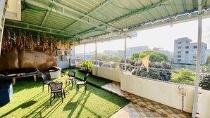 A balcony with artificial grass, chairs, a table, hanging plants, and a view of buildings and greenery. - Hotel One Stay (Indore)