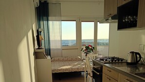 A compact kitchen with a bed in the background, a view of the sea, and a dining area with a table and chairs.