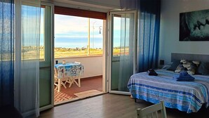 A room with a bed, a table, and a chair. There is a balcony with a view of the beach.
