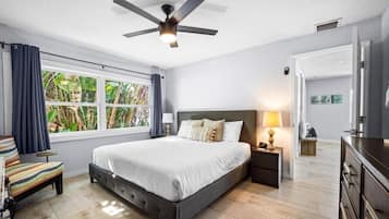 A bedroom with a bed, a nightstand, a lamp, a ceiling fan, a window with curtains, and a view of greenery outside.