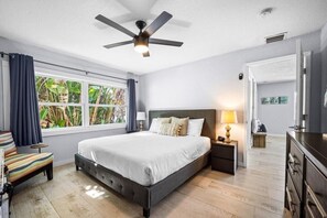 Un dormitorio con cama, mesita de noche, lámpara, ventilador de techo, ventana con cortinas y vistas al exterior.
