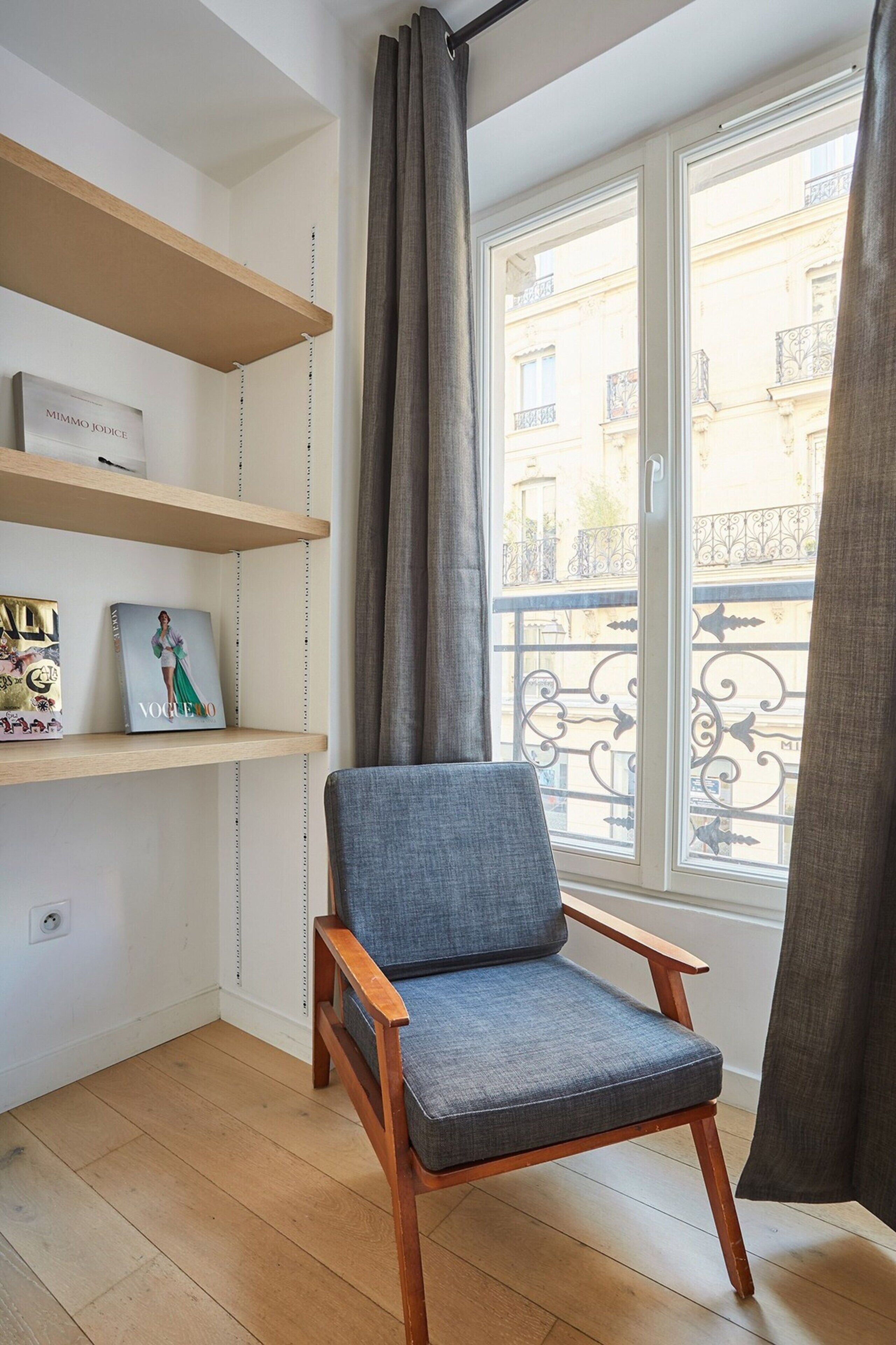 A room with a wooden floor, a grey upholstered chair, a wooden shelf with a Vogue magazine and a framed picture, and a window with a view of buildings and a balcony.
