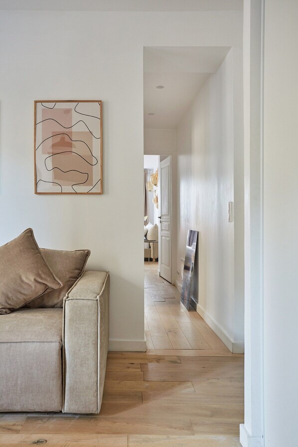 Un salon moderne avec un canapé beige, une œuvre d’art abstraite encadrée et un couloir menant à une autre pièce.