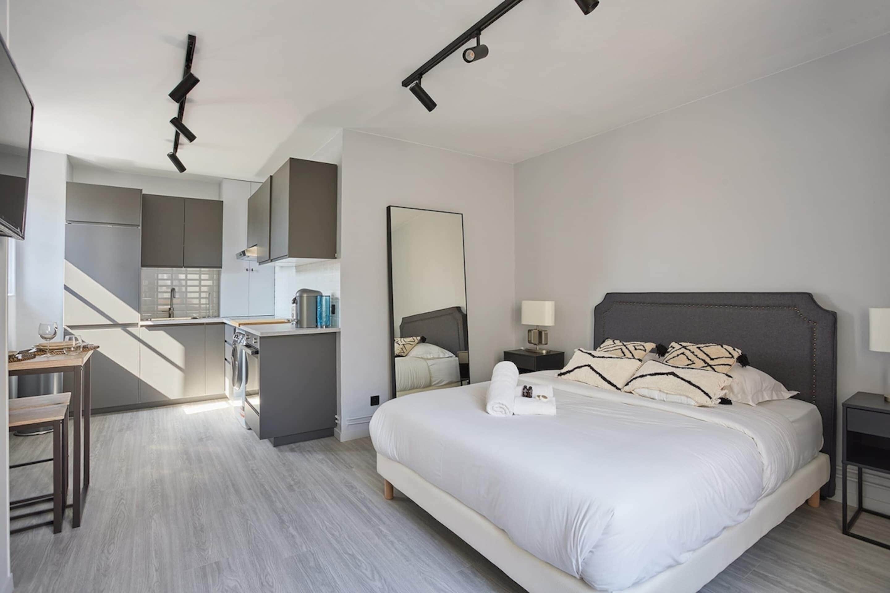 A modern hotel room with a large bed, a desk, a kitchenette, and a bathroom.