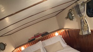 Una cama con ropa de cama blanca y dos almohadas color beige, un cabecero de madera y un pequeño estante con libros y una lámpara.