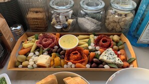 Una tabla de fiambres con diversos quesos, carnes y verduras, acompañada de frascos de pepinillos y aceitunas.