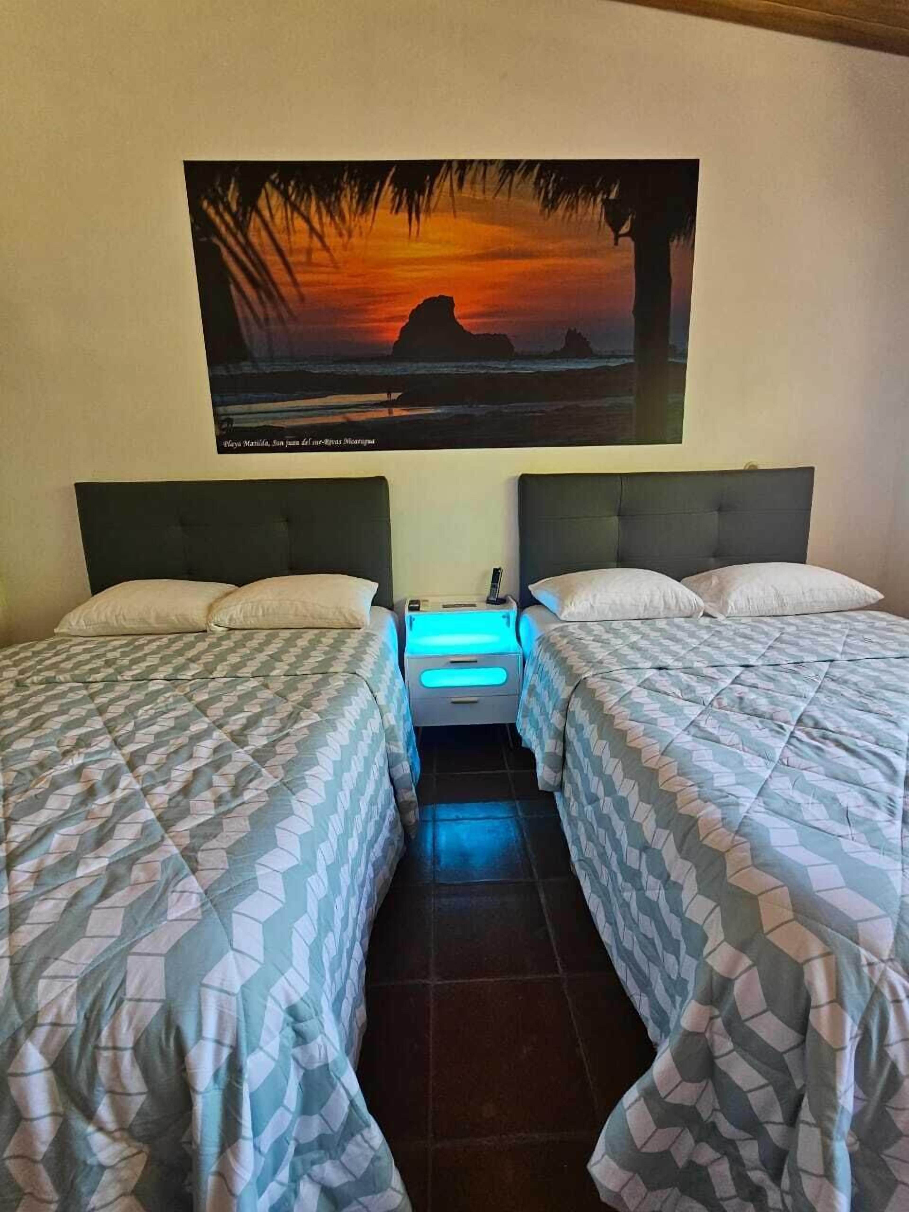 Une chambre avec deux lits, une table de chevet et un tableau représentant un coucher de soleil sur l’océan.