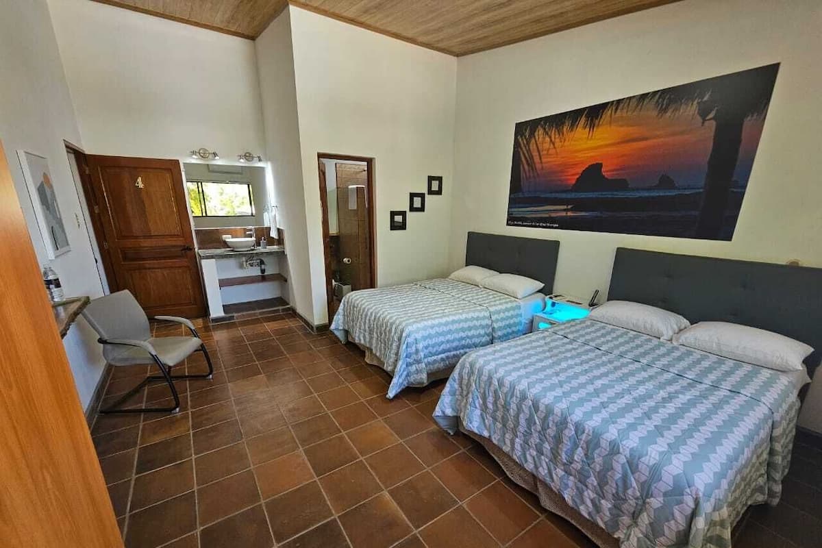 A room with two beds, a desk, and a chair. There is a painting on the wall and a door leading to another room.