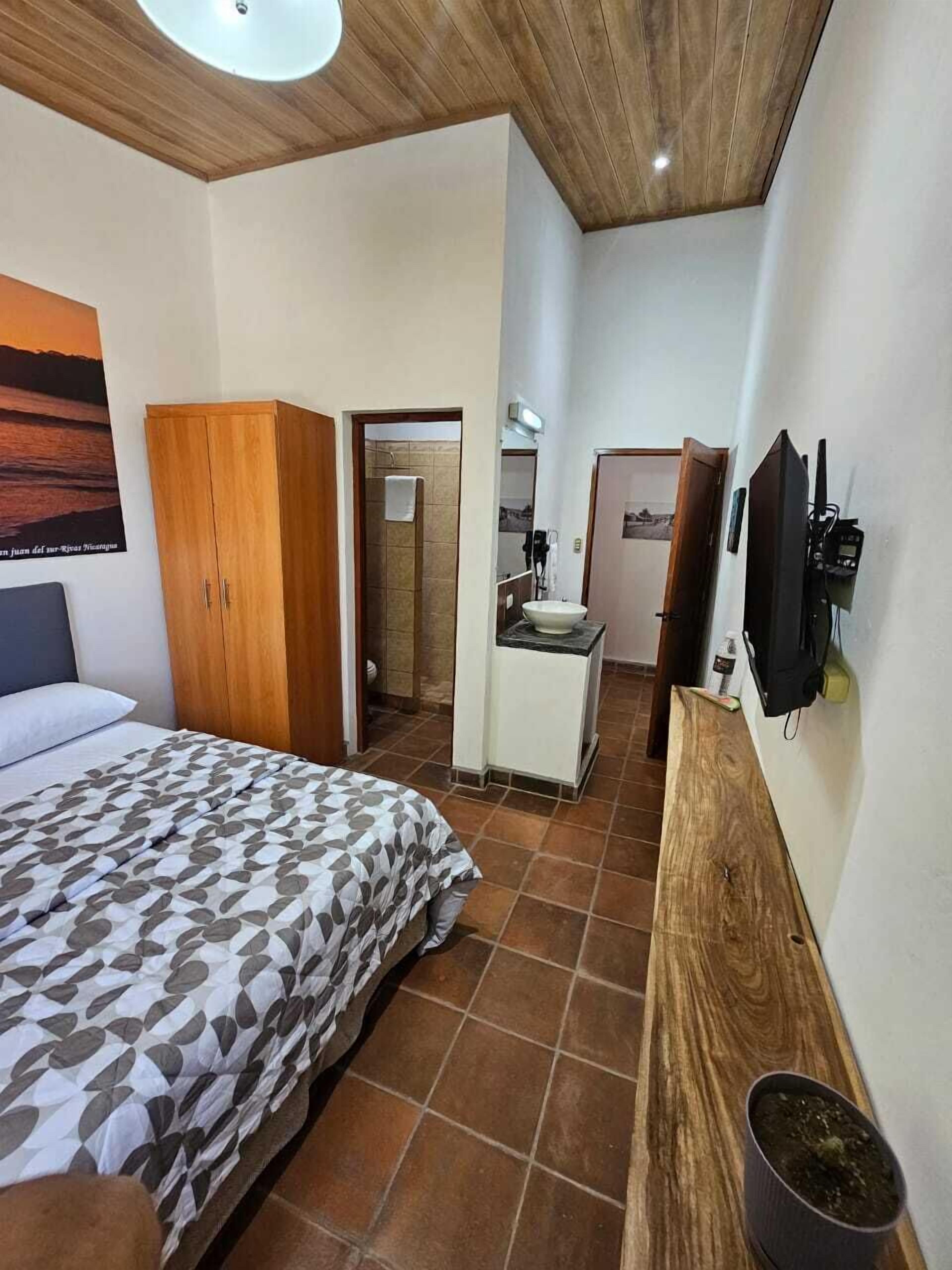 A bedroom with a bed, a wooden wardrobe, a bathroom with a shower, a sink, and a TV mounted on the wall.