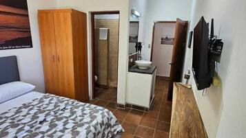 A bedroom with a bed, a wooden wardrobe, a bathroom with a shower, a sink, and a TV mounted on the wall.