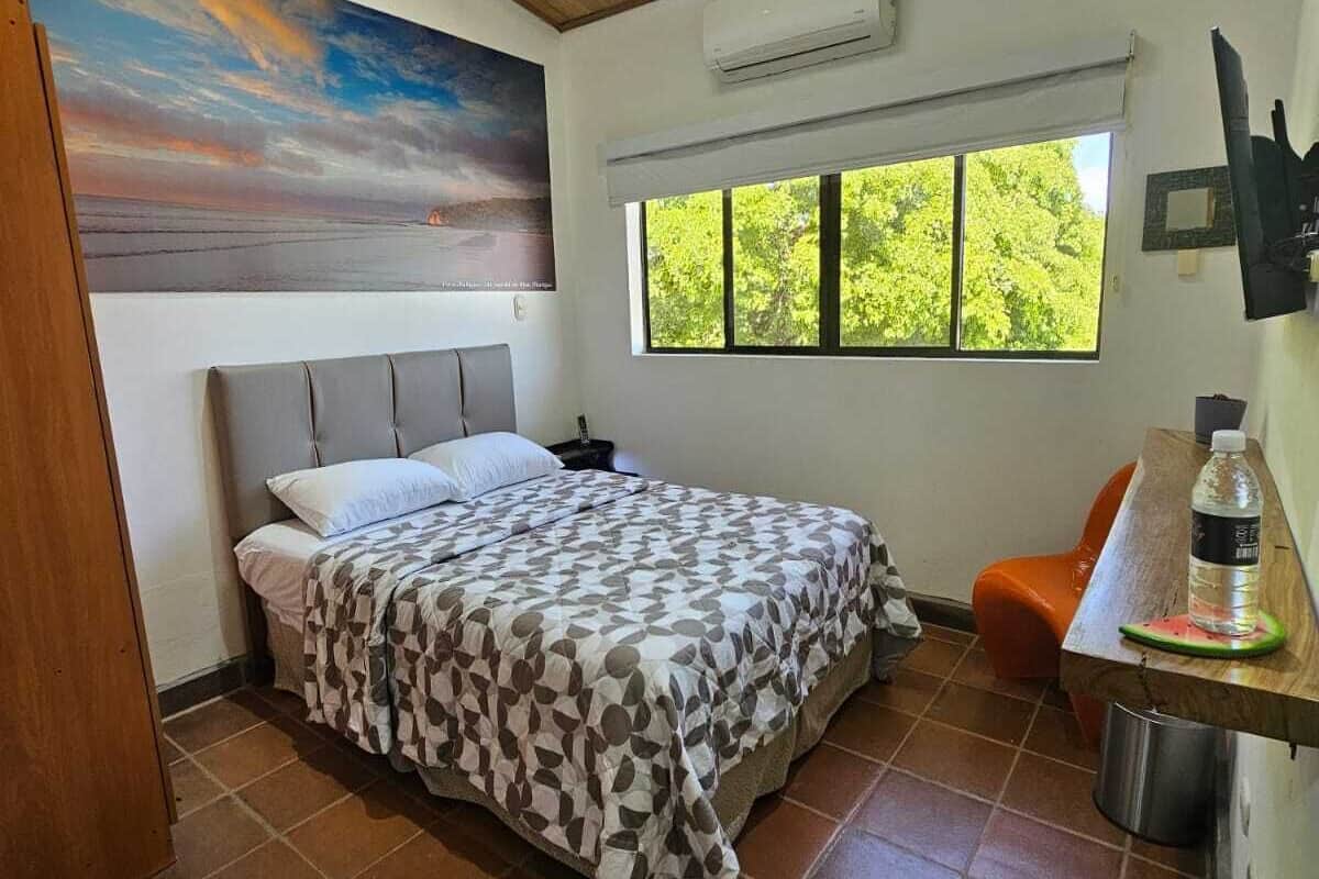 A bedroom with a bed, a painting, a window, and a desk with a water bottle.