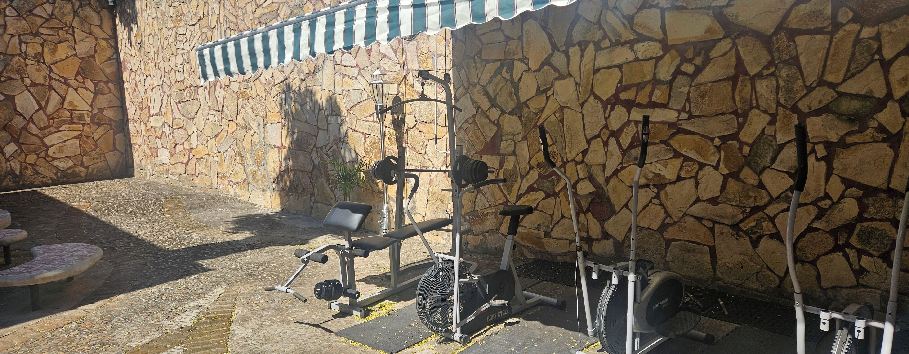 An outdoor gym area with exercise equipment, including a stationary bike and weight machines, against a stone wall backdrop.