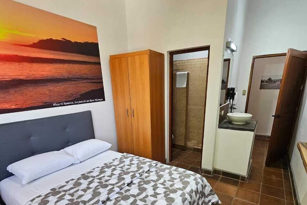 A bedroom with a bed, a wooden wardrobe, a bathroom with a sink and mirror, and a large painting of a sunset.