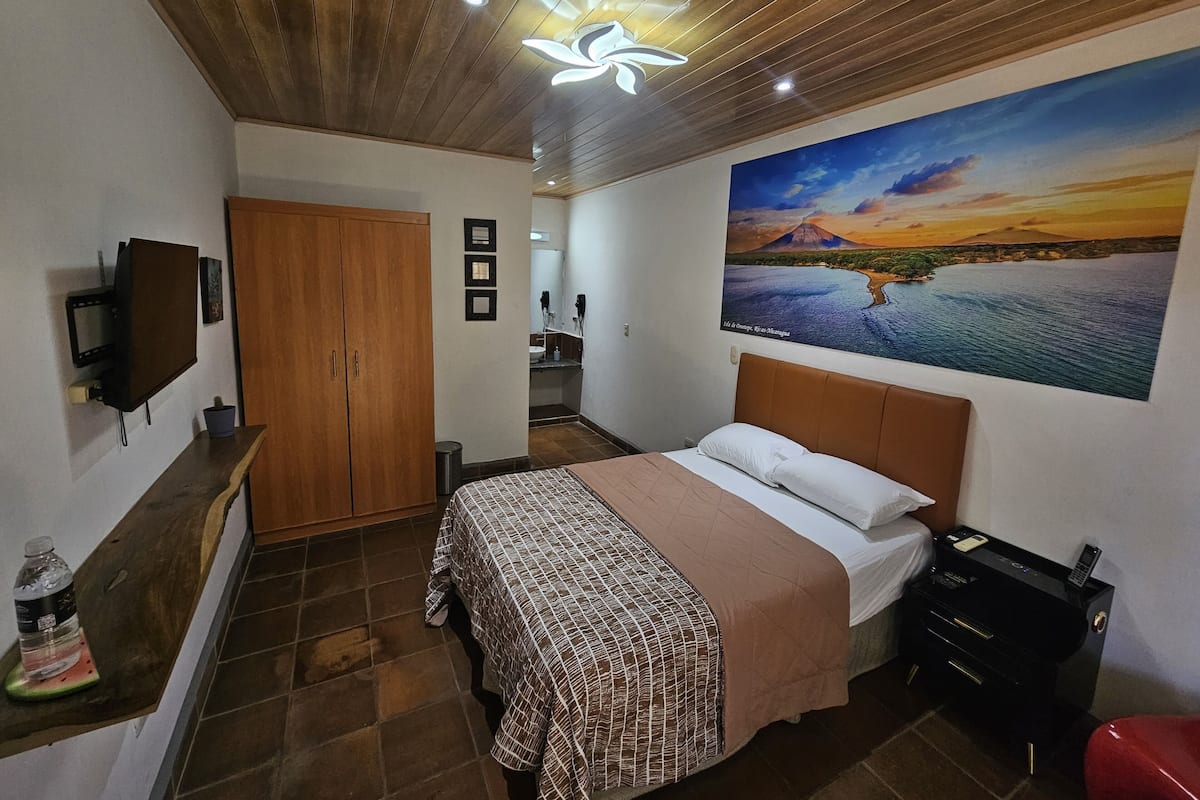 A bedroom with a bed, a wooden wardrobe, a TV, a desk with a printer, and a painting of a landscape.