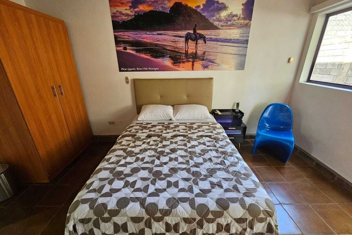 A bedroom with a bed, a wooden wardrobe, a blue chair, and a wall with a sunset painting.