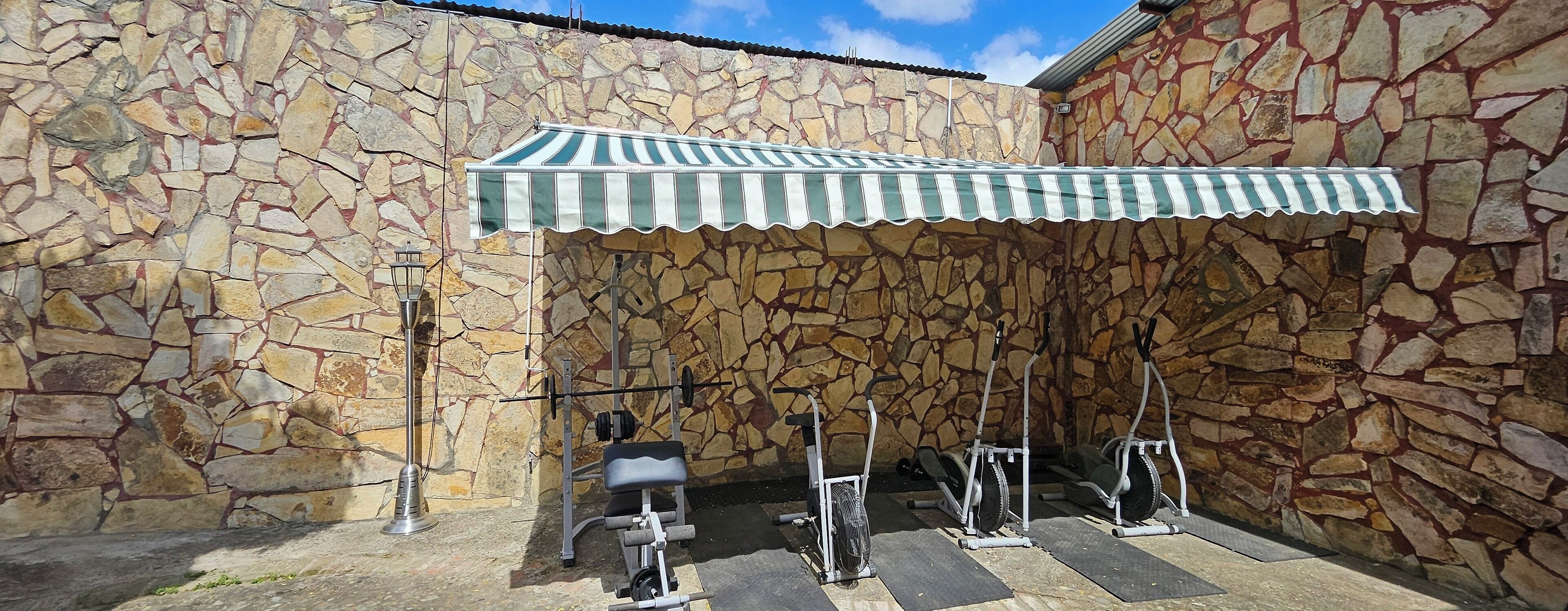 An outdoor gym area with exercise equipment, a stone wall, and a striped awning.