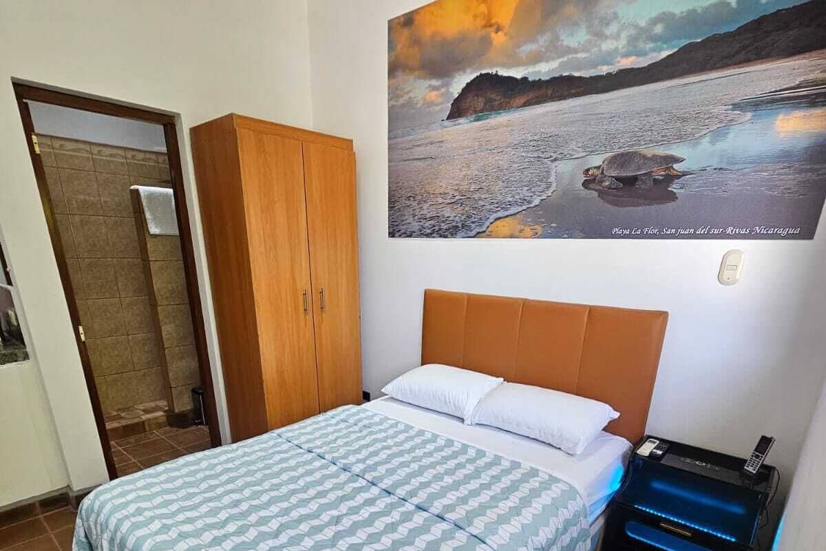 A bedroom with a bed, a wooden wardrobe, a nightstand with a phone, and a wall-mounted painting of a beach scene.