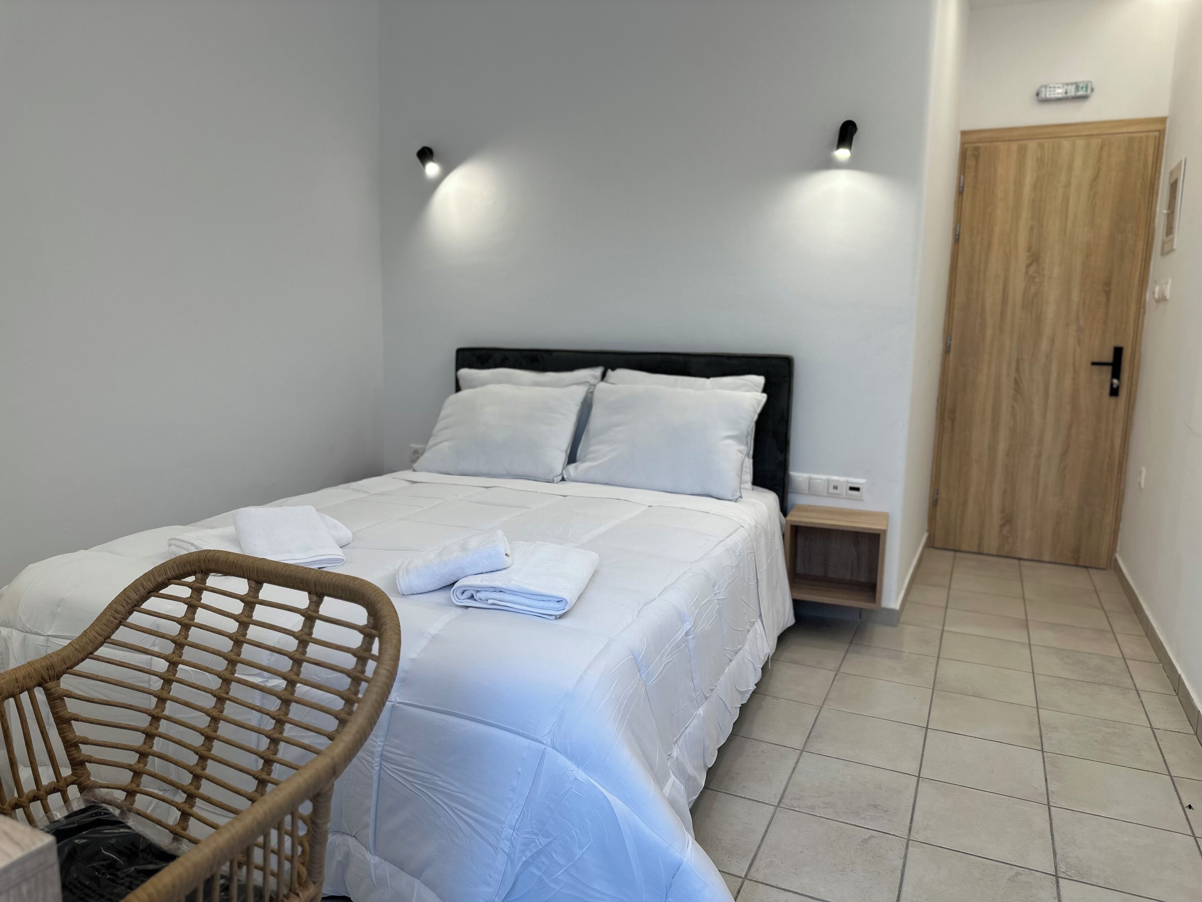 A neatly made bed with white linens, a wooden nightstand, a wicker chair, and a wooden door.