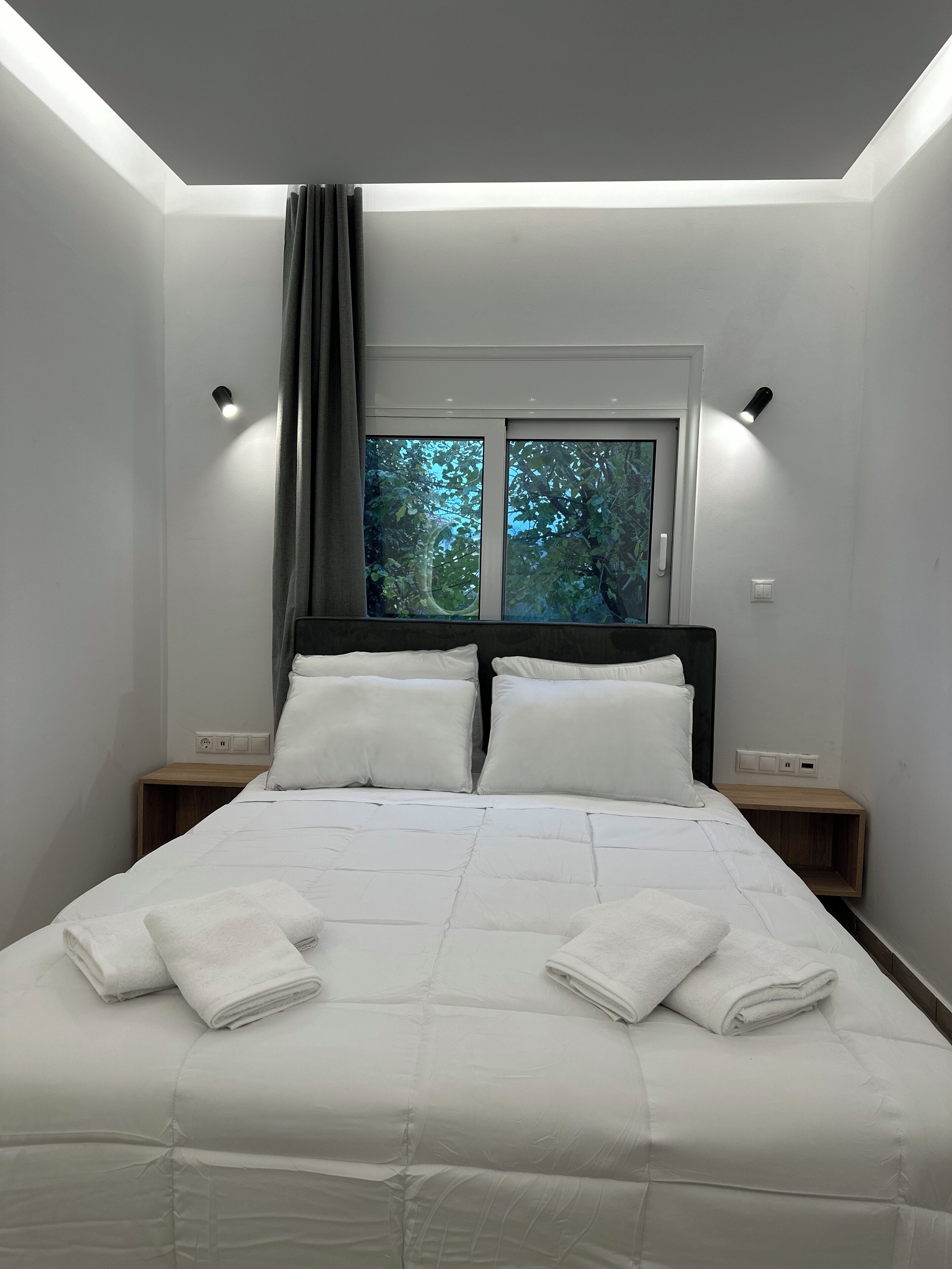A modern bedroom with a large bed, two bedside tables, a window with a view of trees, and a skylight.