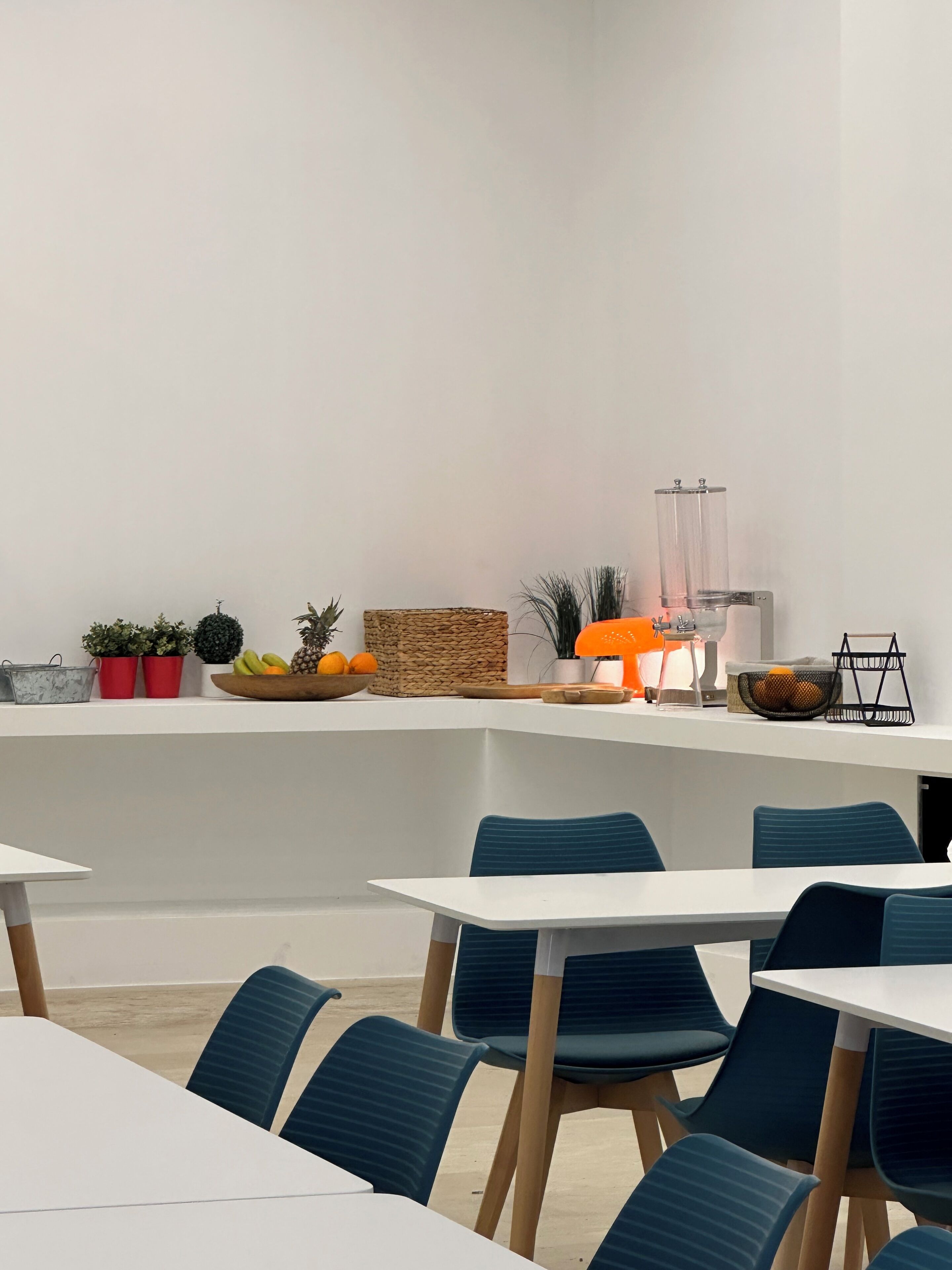 A modern dining area with white tables and blue chairs, a white counter with decorative items, and a clear glass partition.